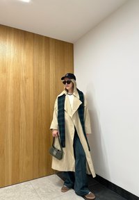 Beige trenchcoat med bred krage, mørkegrønn rutete skjerf, grønn veske og marineblå bukser; bærer solbriller og en svart caps.