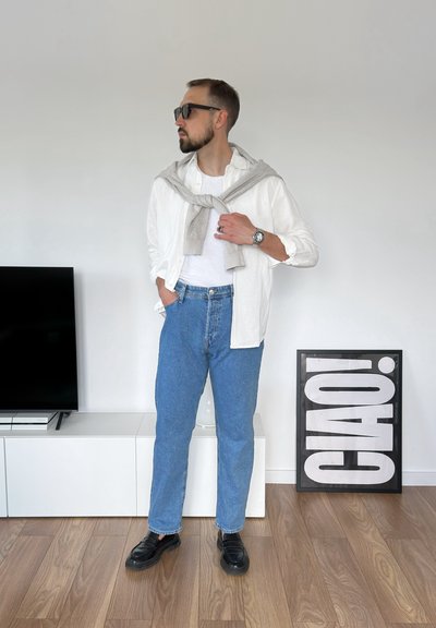 Camisa de lino blanca sobre un suéter gris, combinada con jeans de mezclilla azules y mocasines negros. Presenta gafas de sol y un reloj. Fondo minimalista.