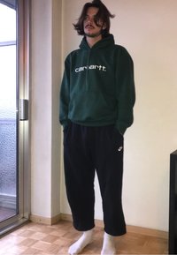 Grøn hættebluse med et hvidt logo, parret med sorte løstsiddende joggingbukser. Står barfodet på en lys trægulv baggrund.