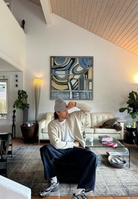 Een hedendaagse woonkamer met een witte leren bank, een glazen salontafel en een houten plafond. Een persoon in een grijze beanie en cardigan zit op een zwarte ottomaan.