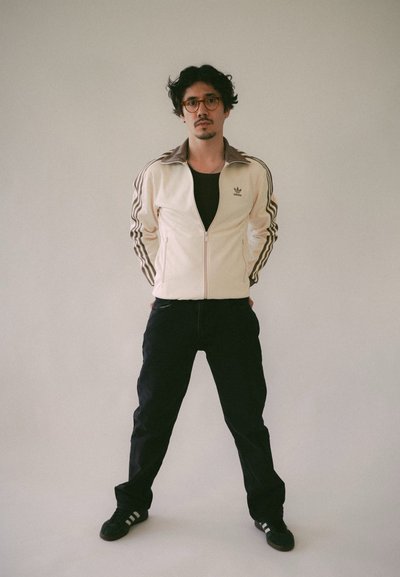 Chaqueta deportiva cremosa con cremallera y rayas oscuras, camiseta negra, pantalones negros y zapatillas negras. Diseño minimalista con logo en la chaqueta.