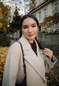 Noor naine tumeda juustega, beežis kohvikus ja pruunis käekotis, seisab lehtedes sügisesel tänaval elamutega lähedal.