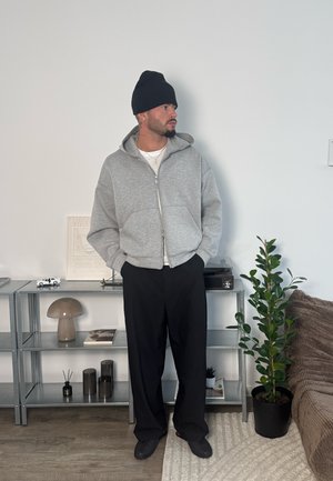 Hombre con gorro negro, sudadera gris con cierre, pantalones negros y zapatos negros, de pie en el interior cerca de estantes de metal y una planta en maceta.