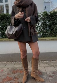 Brūns ādas rokassomu, liela izmēra melna žakete, brūna šalle ar bārkstīm, pleatota melna svārku, un tan krāsas ceļgala augstiem zābakiem uz bruģētas virsmas.