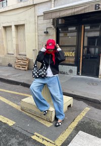 Personne portant une casquette rouge, une veste noire, un jean large bleu clair et des chaussures à pois, assise sur un piédestal de rue, ajustant sa casquette et tenant un sac noir à pois.