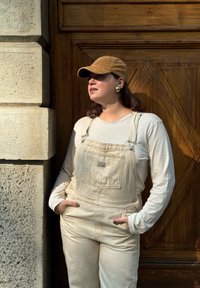 Salopette en denim beige avec poche frontale et quincaillerie argentée, superposée sur une chemise à manches longues légère. Casquette en velours côtelé marron et grandes boucles d'oreilles.