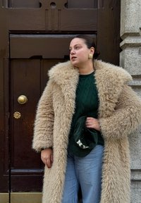 Cappotto in pelliccia sintetica beige con ampio colletto, maglione lavorato a trecce verde scuro, denim azzurro chiaro e cappello verde scuro con scritte bianche.