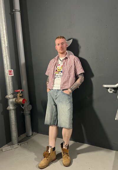 Camisa de cuadros roja y blanca de manga corta sobre una camiseta gráfica, pantalones cortos de mezclilla y botas marrones con acentos de piel, de pie contra una pared oscura.