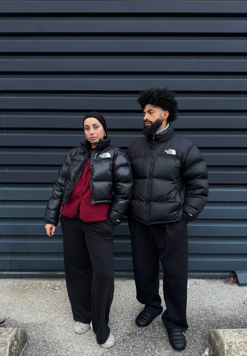 Deux personnes portant des doudounes noires se tiennent devant un mur métallique gris foncé, l'une les mains dans les poches, l'autre le bras détendu.