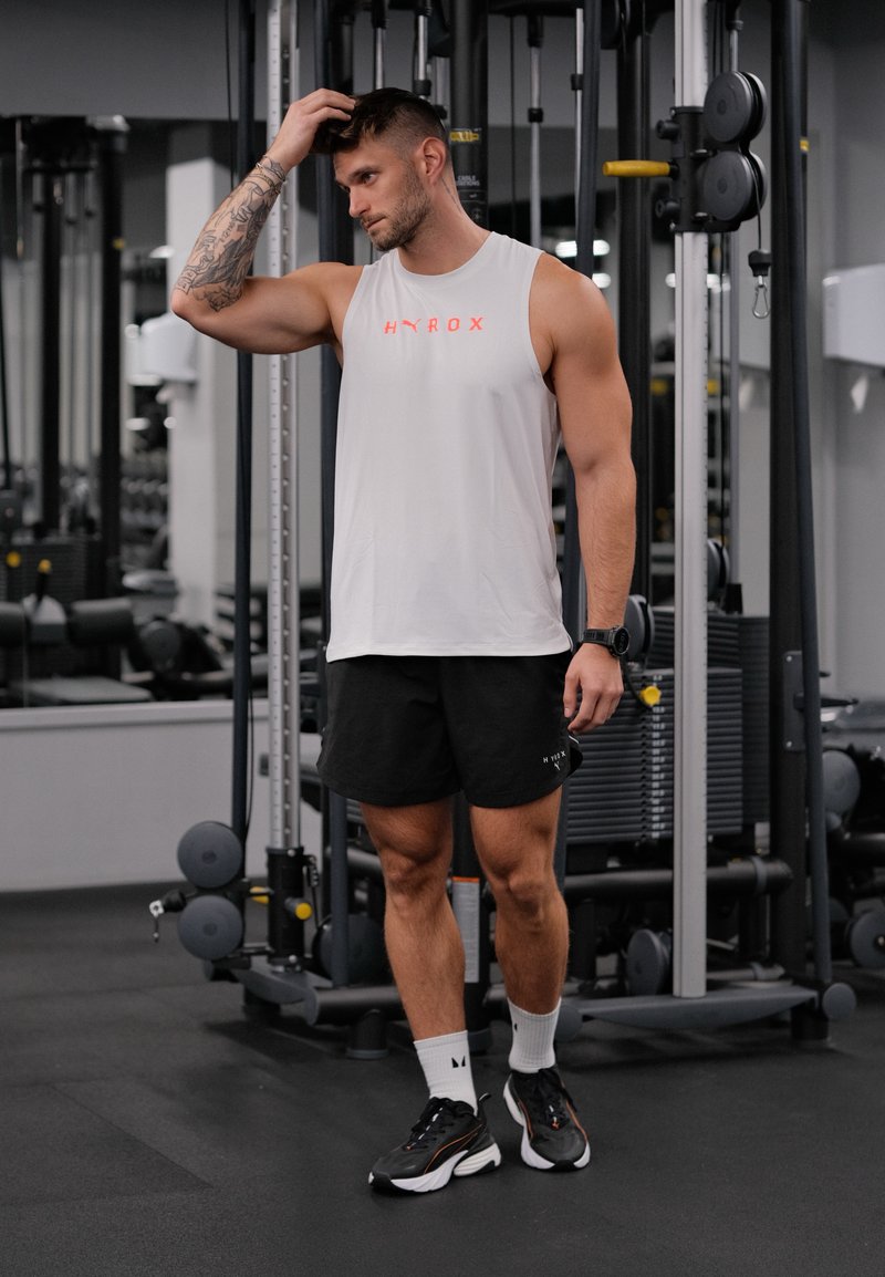 Chemise sans manches gris clair avec logo rouge, shorts noirs, chaussures de sport noires et chaussettes blanches. En arrière-plan, équipement de gym.