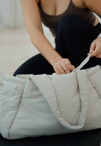 Personne ouvrant un sac matelassé beige clair en étant à genoux, portant un haut marron sans manches et un pantalon noir.