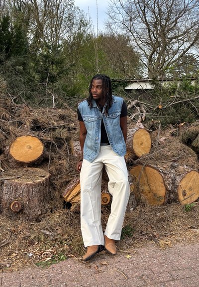 Chaleco de mezclilla sobre una camisa negra, acompañado de pantalones claros y zapatos marrones. El fondo presenta troncos cortados y vegetación escasa.