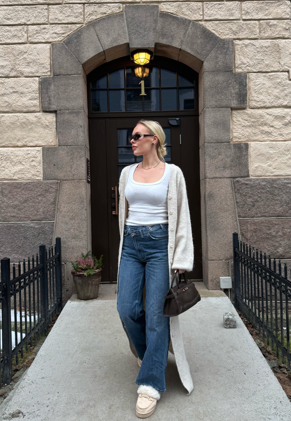 Femme marchant sur un chemin à l'extérieur d'un bâtiment en pierre avec une porte voûtée sombre numérotée 1, portant un débardeur blanc, un long cardigan, un jean, des lunettes de soleil et tenant un sac marron.