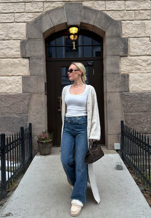 Mujer caminando por un camino frente a un edificio de piedra con una puerta arqueada y oscura numerada con el 1, vistiendo una camiseta blanca sin mangas, un largo cárdigan, vaqueros, gafas de sol y sosteniendo un bolso marrón.
