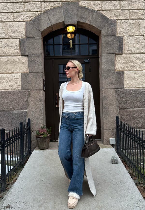 Vrouw die op een pad loopt vóór een stenen gebouw met een donkere boogvormige deur met nummer 1, gekleed in een wit hemdje, een lange cardigan, jeans, zonnebril en een bruine tas vasthoudend.