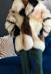 Personne portant un manteau en fourrure duveteux multicolore avec un pantalon vert, debout près d'un canapé bleu avec un coussin à motifs zèbre.