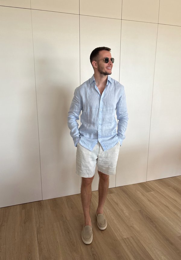 Mann trägt hellblaues Langarmhemd, weiße Shorts, beigen Slip-on Schuhe und Sonnenbrille, steht vor einer schlichten Wand auf Holzboden.