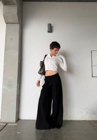 Jeune personne aux cheveux courts et bouclés portant un haut court blanc et un pantalon large noir, posant contre un mur blanc avec un sac sur une épaule.