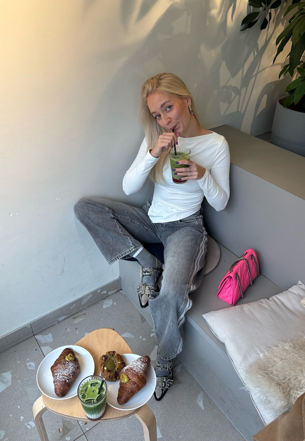 Dva rogljička na belih krožnikih s sladkorjem v prahu, zelena pijača v kozarcu, roza torbica ter oseba v beli majici z dolgimi rokavi.