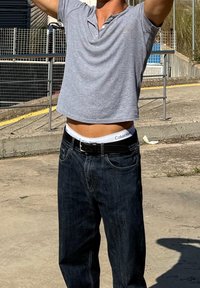 Grå kortærmet polo, mørke jeans, sort bælte, synlig Calvin Klein underbukse, udendørs omgivelser med en betonoverflade.
