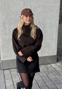 Brun oversized tröja med hög krage, bärs över en svart kjol. Accessoarer med en brun keps. Slät textur, minimal design.