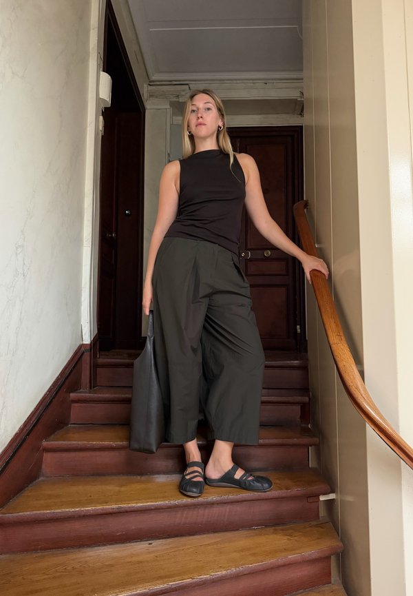 Mujer de pie en unas escaleras de madera sosteniendo un bolso negro, vistiendo una camiseta negra sin mangas, pantalones anchos negros y zapatos Mary Jane negros.