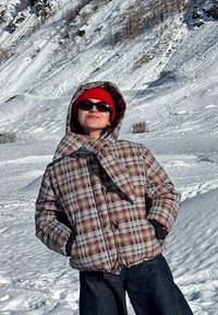 Personne portant une veste d'hiver à carreaux, un bonnet rouge et des lunettes de soleil, debout les mains dans les poches dans un paysage de montagnes enneigées.