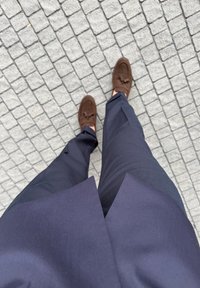 Sötétkék nadrágot és barna mokaszint viselő személy áll szürke macskaköves járdán.