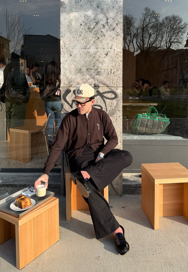 Uomo con cappello e occhiali seduto fuori da un bar su sgabelli di legno, che porge la mano verso un bicchiere di latte vicino a un dolce su un vassoio.