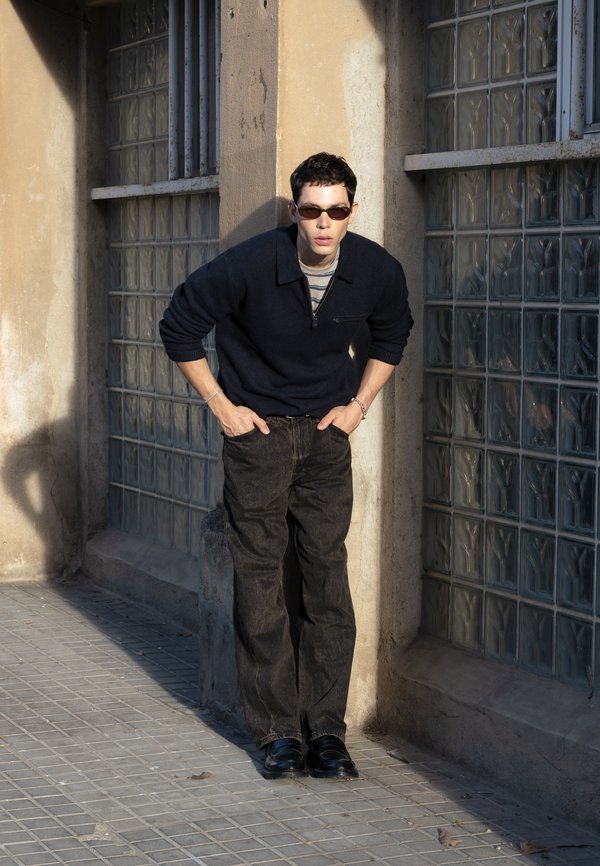 Joven con gafas de sol, suéter negro y pantalones oscuros se inclina hacia adelante con las manos en los bolsillos, de pie junto a un edificio con ventanas de bloques de vidrio.