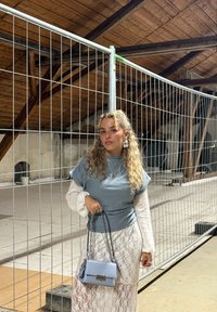Mladá žena s kučeravými vlasmi stojí vo vnútri pri kovovej plote, oblečená v vrstvenom šedom a bielom oblečení a drží svetlomodrú kabelku s retiazkou.
