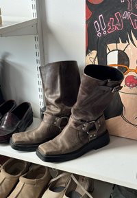 Bottes mi-mollet en cuir brun avec boucles sur une étagère blanche à côté de diverses chaussures et d'une peinture colorée de style cartoon.