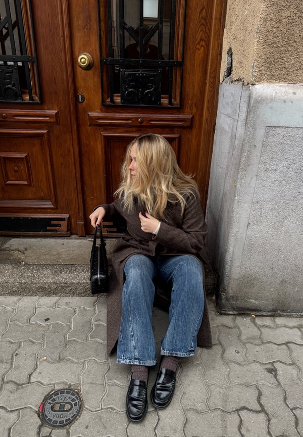 Valkā garu brūnu mēteļu, zilas izplešanās džinsas un melnas mokasīnus. Melna rokassoma atrodas blakus personai, kas sēž pie koka durvīm.