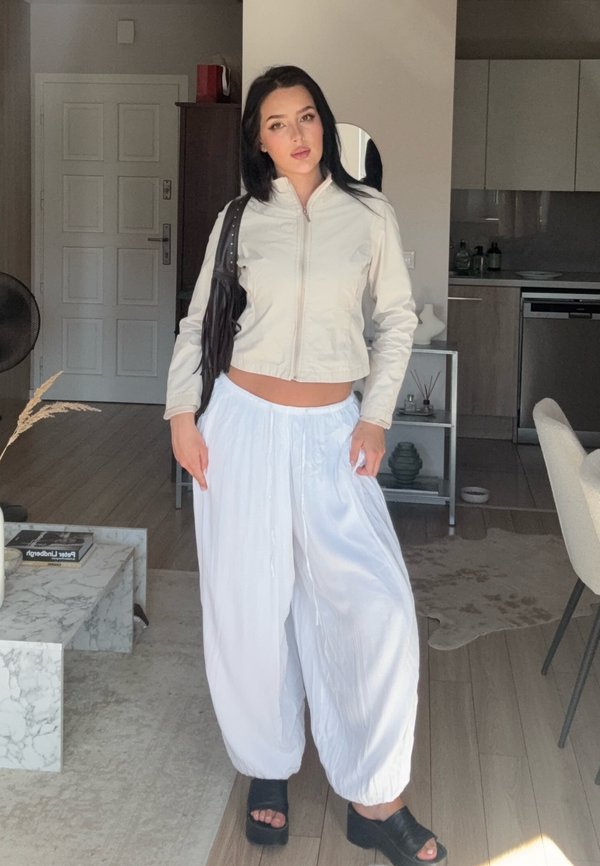 Mujer de pie en el interior con una chaqueta corta ligera, pantalones blancos holgados, sandalias negras de plataforma y lleva un bolso de hombro negro con flecos.
