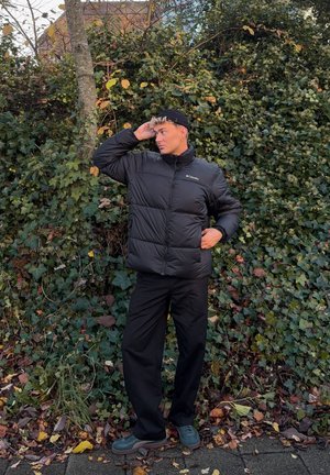 Chaqueta de plumas negra con un alto cuello, que presenta un diseño acolchado. Combinada con pantalones oscuros y zapatos verdes, con un fondo de vegetación.