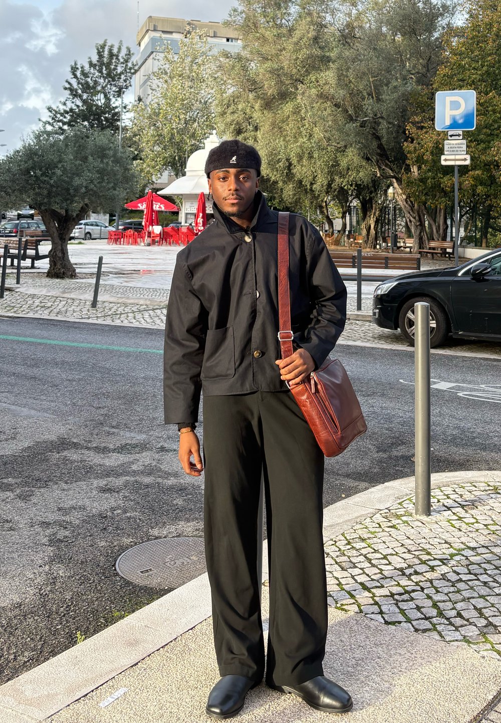 Giovane uomo con giacca e pantaloni neri e borsa marrone a tracolla si trova in una strada della città vicino a un'auto parcheggiata e tavoli di un bar all'aperto.