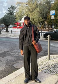 Giovane uomo con giacca e pantaloni neri e borsa marrone a tracolla si trova in una strada della città vicino a un'auto parcheggiata e tavoli di un bar all'aperto.