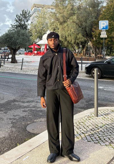 Joven vestido con chaqueta y pantalones negros, con un bolso marrón al hombro, está de pie en la calle de la ciudad cerca de un coche estacionado y mesas de un café al aire libre.