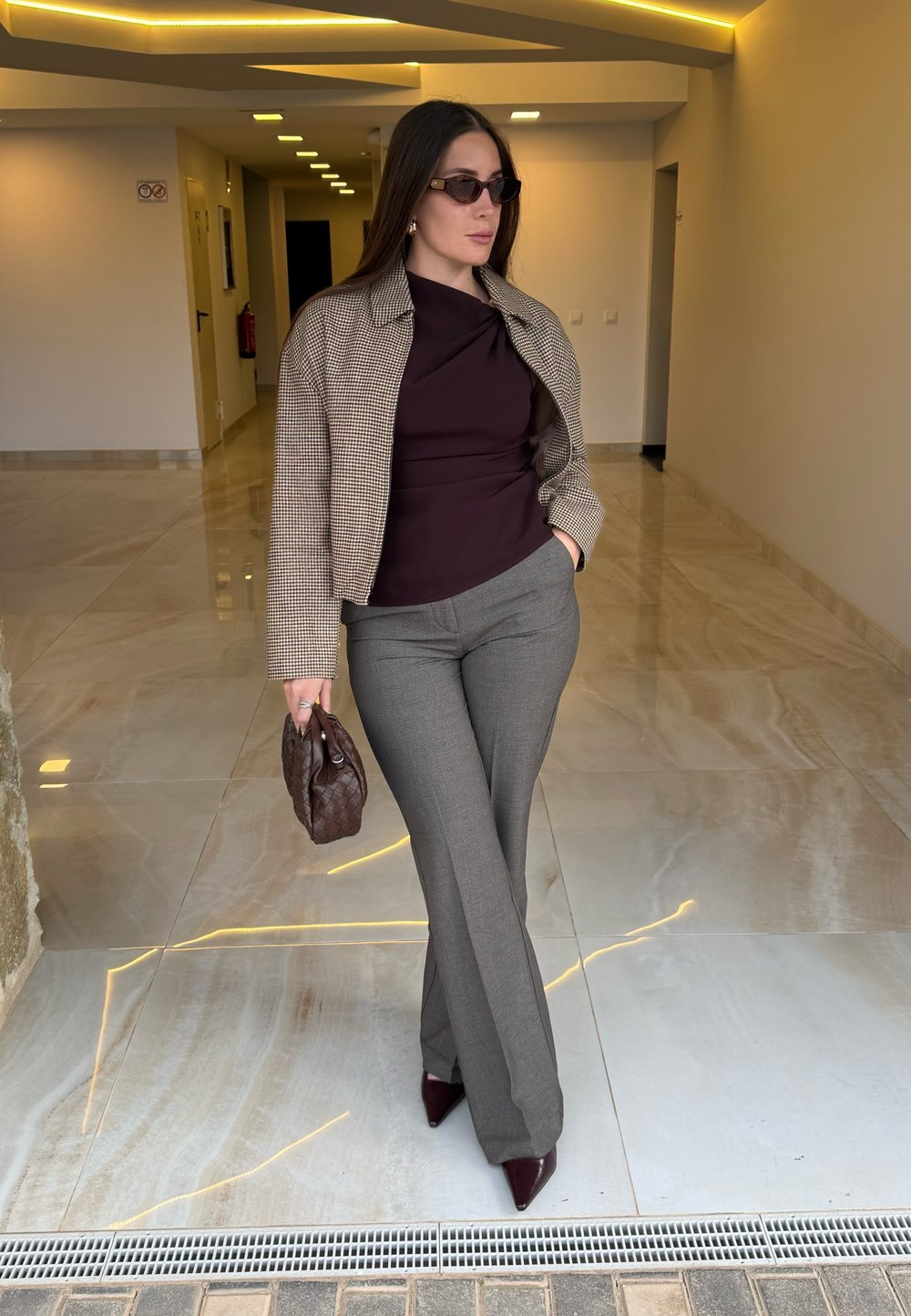 Veste courte à carreaux marron et beige portée sur un haut mauve foncé, assortie à un pantalon gris évasé et des escarpins pointus rouge foncé. Tenant un sac marron.