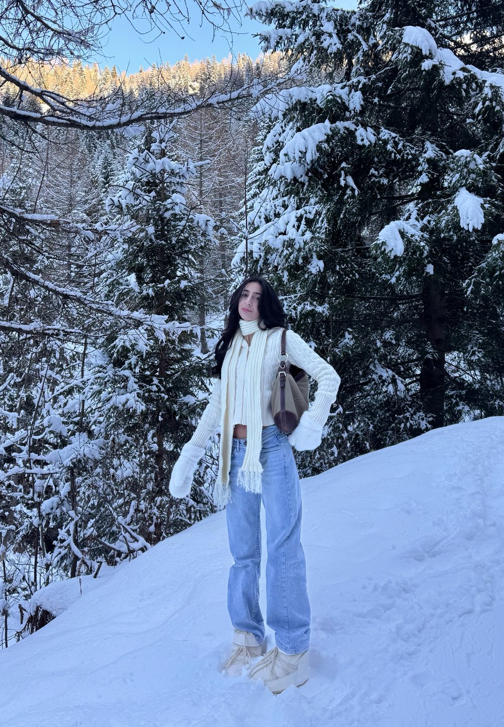 Femme portant une écharpe blanche, des moufles, un pull et un jean debout sur une pente enneigée avec des arbres à feuilles persistantes chargés de neige en arrière-plan.