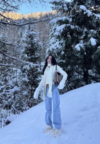 Femme portant une écharpe blanche, des moufles, un pull et un jean debout sur une pente enneigée avec des arbres à feuilles persistantes chargés de neige en arrière-plan.