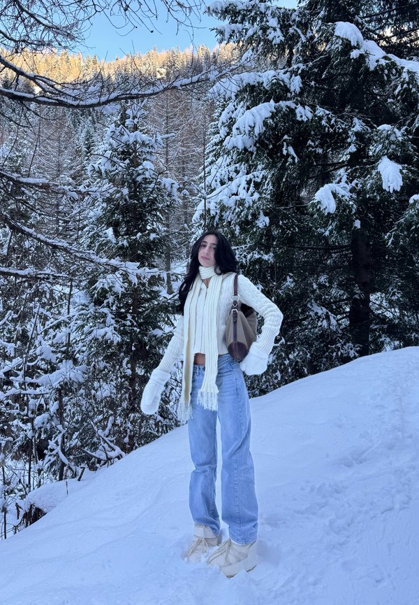 Nő fehér sállal, kesztyűvel, pulóverrel és farmernadrággal, hóval borított lejtőn áll, háttérben havas örökzöld fákkal.