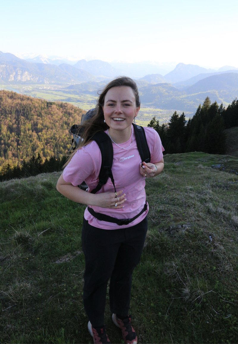 Femme souriante en chemise rose et pantalon noir ajuste son sac à dos tout en se tenant sur une colline herbeuse avec une forêt et des montagnes en arrière-plan.