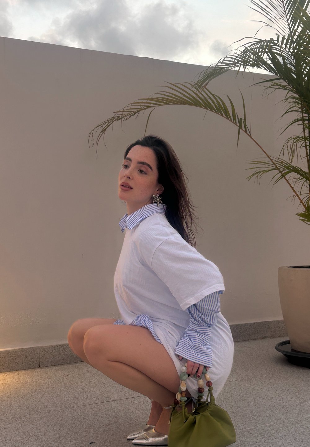 Bílé oversized tričko s modrým pruhovaným límečkem, spárované se stříbrnými slip-on botami a zelenou taškou se zdobnými korálky. Světlé pozadí, palma.