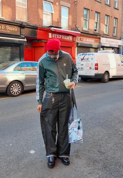 Chaqueta de chándal verde de Adidas con rayas blancas, pantalones de mezclilla oscuros holgados, zapatos negros y una gorra de béisbol roja. Lleva una bolsa transparente con ropa.