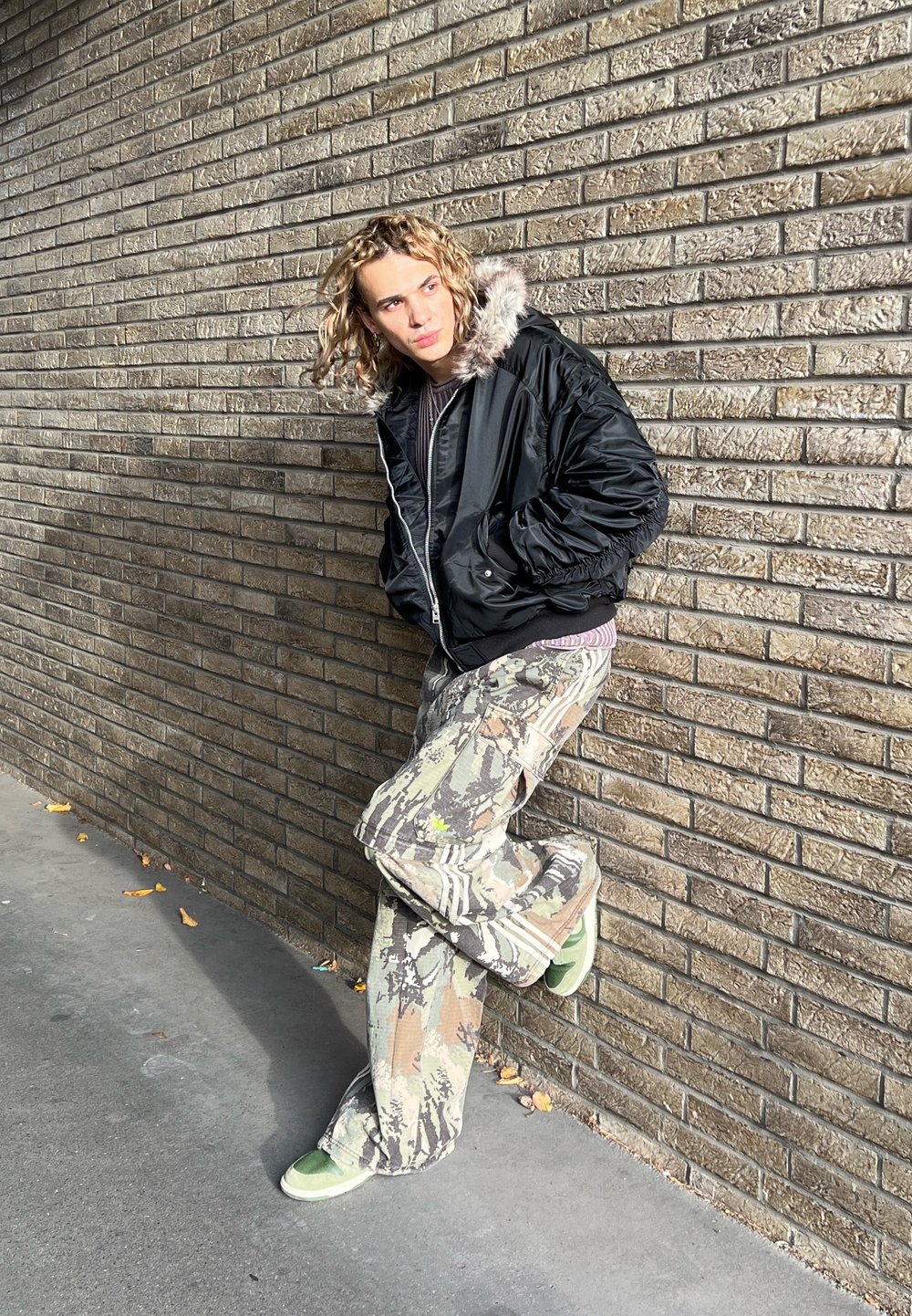 Sort bomberjakke med pelsforet hætte, kombineret med camouflage baggy bukser i nuancer af beige, grøn og brun. Grønne sko på beton.