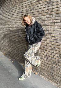 Sort bomberjakke med pelsforet hætte, kombineret med camouflage baggy bukser i nuancer af beige, grøn og brun. Grønne sko på beton.