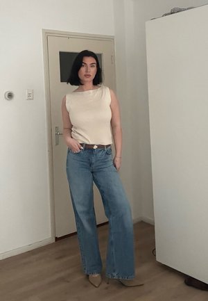 Mujer con cabello negro, vestida con top beige sin mangas, jeans azules de pierna ancha y tacones beige puntiagudos, de pie en el interior junto a una puerta y un armario blanco.