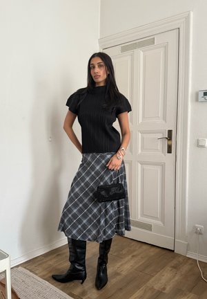 Mujer con blusa negra de manga corta, falda midi a cuadros gris, botas negras hasta la rodilla, sosteniendo un bolso pequeño negro, de pie en el interior cerca de una puerta blanca.