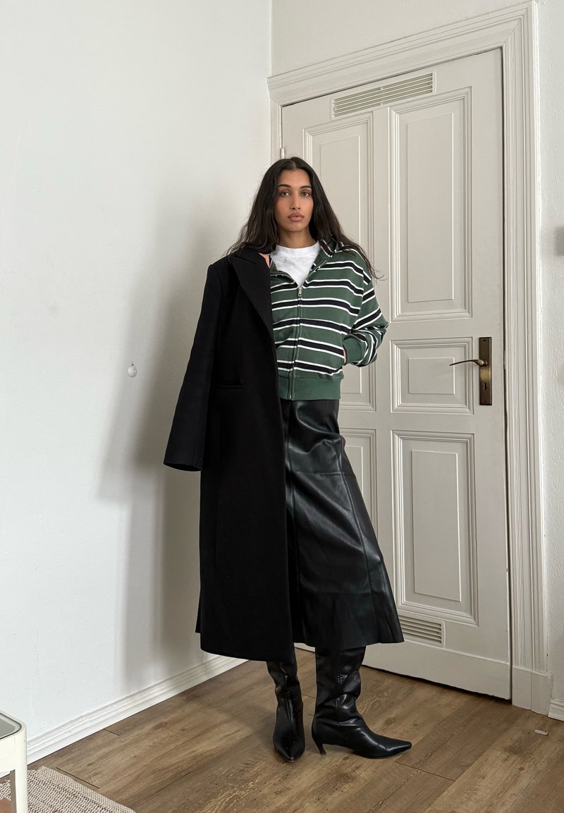 Femme aux longs cheveux foncés portant un sweat à capuche zippé vert rayé, une jupe en cuir noire, des bottes noires et un manteau noir posé sur une épaule à l'intérieur.